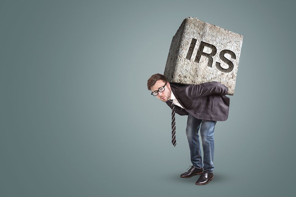 A man in a suit and glasses struggles to carry a large concrete block labeled "IRS" on his back, set against a plain gradient background.