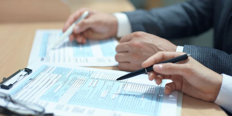Two businesspeople in suits sign documents at a table, with pens in hand and forms in front of them.