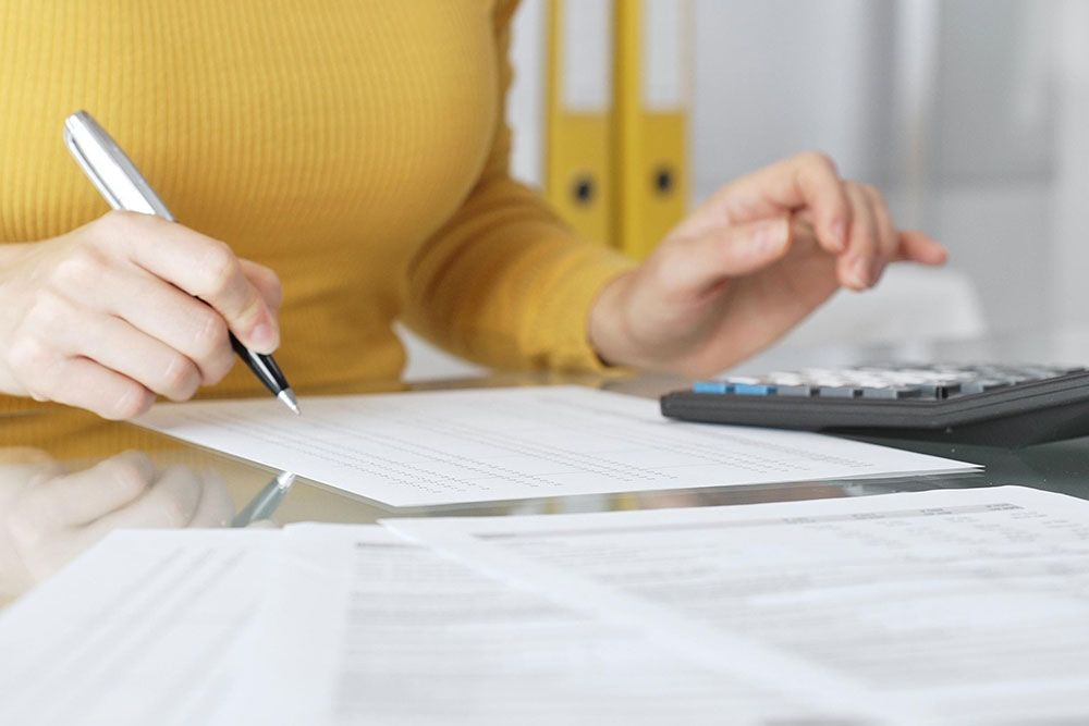 A person wearing a yellow sweater reviews financial documents, holding a pen and using a calculator, seated at a desk with additional papers.