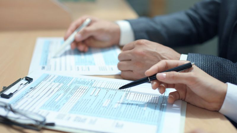 Two businesspeople in suits sign documents at a table, with pens in hand and forms in front of them.