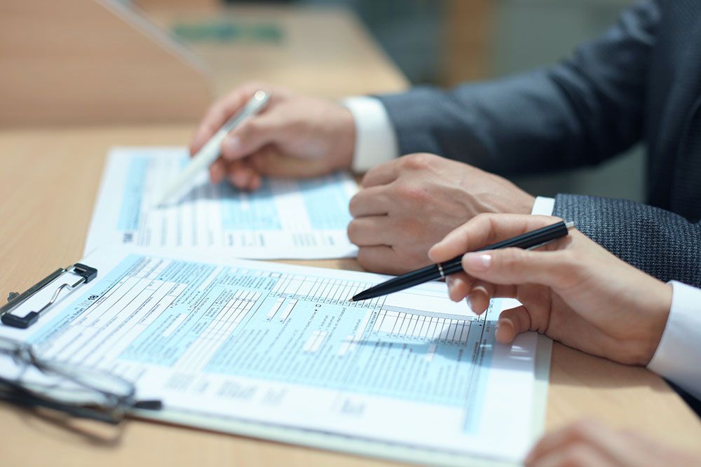 Two businesspeople in suits sign documents at a table, with pens in hand and forms in front of them.