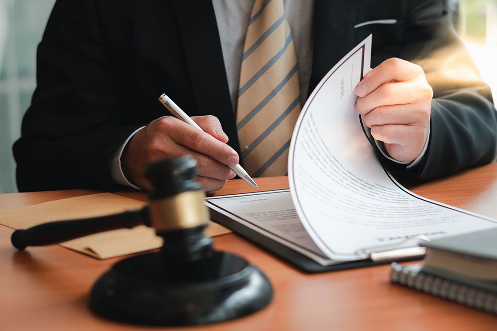 Close-up of a person in a suit signing documents at a desk, with a gavel in the foreground.