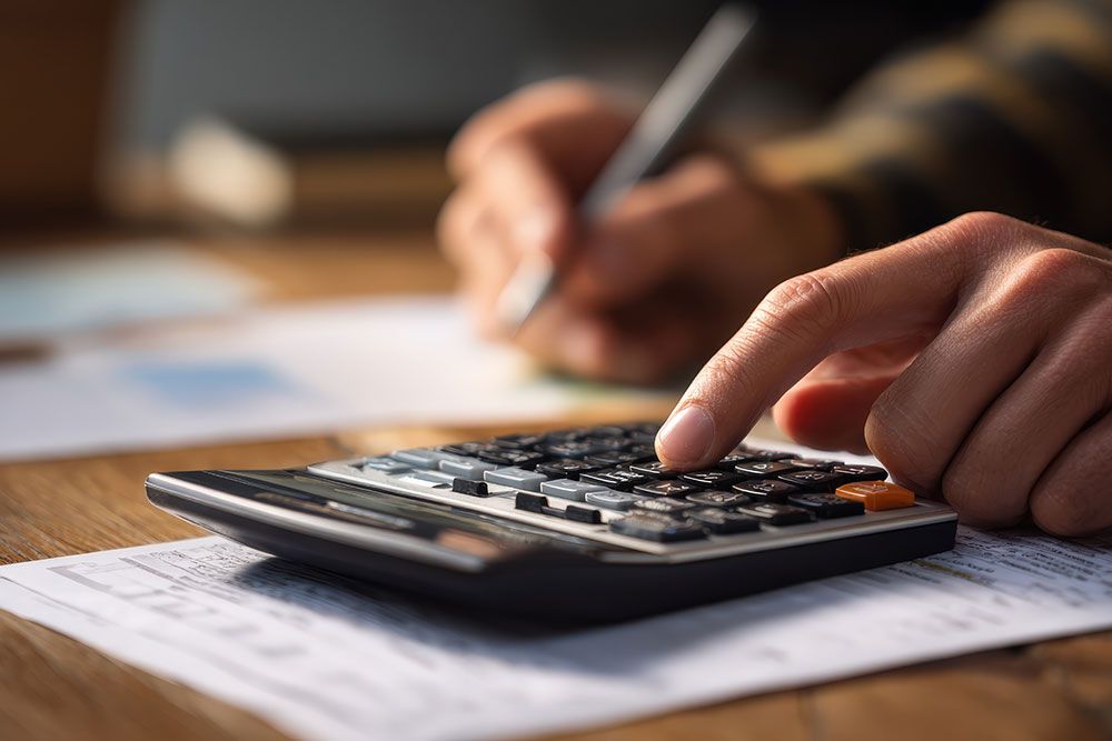 Hand pressing a calculator button on a wooden desk, with the other hand writing on scattered paper documents, indicating a focus on calculations and note-taking.