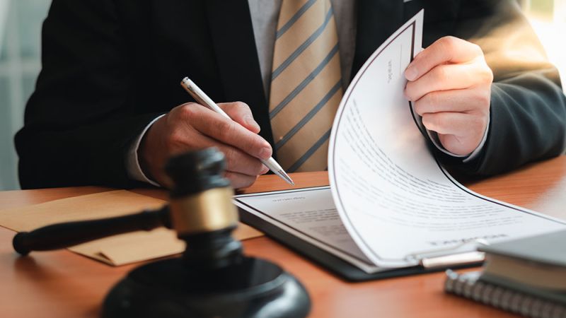 Close-up of a person in a suit signing documents at a desk, with a gavel in the foreground.